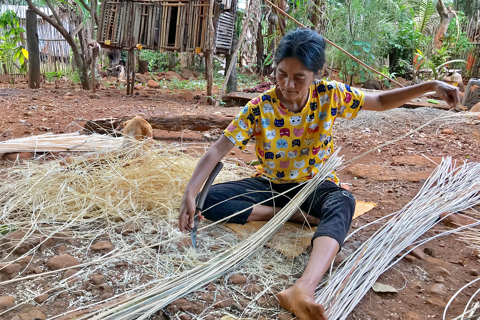 A woman thinning rattan strips in the shade of a tree.